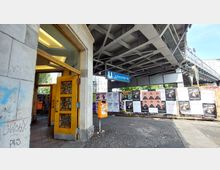 Eingangsbereich einer U-Bahn-Station an der Haltestelle "Schlesisches Tor" mit einer geöffneten Holztür und verzierten Glasscheiben. Im Außenbereich sind eine Metallkonstruktion der Hochbahn, zahlreiche Plakate an einer Wand und ein oranger Mülleimer sichtbar.