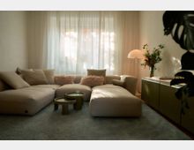 Wohnzimmer mit einem großen beigefarbenen Sofa, dekoriert mit flauschigen Kissen, einem flauschigen Teppich und einem Satz runder Couchtische. Im Hintergrund befinden sich bodenlange, helle Vorhänge vor einem Fenster, eine Stehlampe mit rundem Schirm und eine Anrichte mit einer Vase mit Blumen.