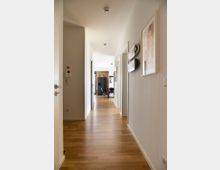 Langer, heller Flur in einer modernen Wohnung mit Holzboden und weißen Wänden. An den Wänden hängen Bilder und Dekorationen, und am Ende des Flurs ist ein Essbereich mit einem Tisch und Stühlen sichtbar.