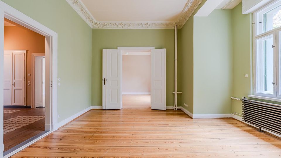 Heller, leerer Altbau-Wohnraum mit hellgrünen Wänden, breiten Dielenböden und Stuckdecke. Weiße Doppeltüren führen in angrenzende Räume, rechts sorgt ein großes Fenster mit Heizkörper darunter für Tageslicht.