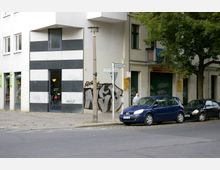 Straßenecke mit einem modernen Gebäude, dessen Fassade abwechselnd aus schwarzen und weißen horizontalen Streifen besteht. Im Vordergrund stehen zwei geparkte Autos, während Straßenschilder und ein Baum die Umgebung definieren; Graffiti sind auf einer angrenzenden Wand sichtbar.