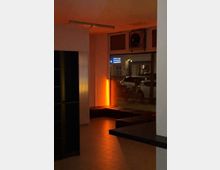 Innenraum eines kleinen Eingangs- oder Wartebereichs mit gefliester Bodenfläche, gepolsterten Sitzbänken und warmem orangefarbenem Licht. Eine große Fensterfront zeigt den Blick auf die Straße mit geparkten Autos und Fahrrädern; darüber sind Lüftungselemente und eine Deckenleuchte zu sehen.