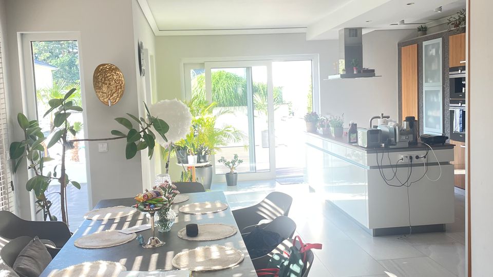 Moderner, heller Ess- und Wohnbereich mit einem großen Esstisch, umgeben von Stühlen, dekoriert mit Platzsets und Blumen. Im Hintergrund eine offene Küche mit weißen Fronten und große Fenster, die zu einer Terrasse mit Pflanzen führen.