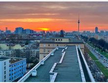 Städtische Ansicht von Berlin bei Sonnenuntergang, mit der Silhouette des Berliner Fernsehturms im Hintergrund. Im Vordergrund sind Gebäude und Straßen zu sehen, die von Grünflächen und Baumreihen gesäumt werden.