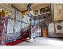 Prunkvolles Treppenhaus mit verziertem roten Marmorgeländer, einer roten Teppichtreppe und gemusterten Marmorwänden. Goldene Verzierungen, ein großer Kerzenleuchter und ein prächtiges Holzportal ergänzen die luxuriösen Details, während an der Decke und Wänden dekorative Gemälde zu sehen sind.