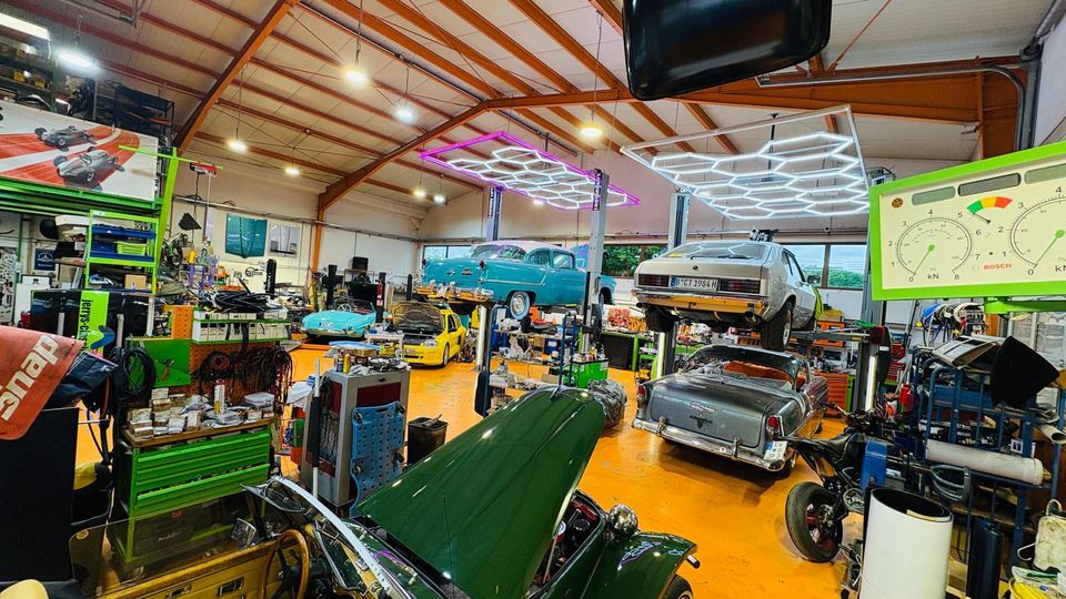Große Autowerkstatt mit hoher Hallendecke und orangefarbenem Boden, voll mit Werkzeugwagen, Regalen und Ersatzteilen. Mehrere Oldtimer stehen auf Hebebühnen, darüber hängen helle, sechseckige LED-Leuchten und eine große Anzeigeinstrument-Tafel ist im Vordergrund zu sehen.