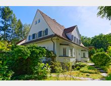 Ein zweistöckiges Haus mit weißer Fassade und steilem Walmdach, umgeben von einem gepflegten Garten mit Sträuchern, Bäumen und einer Steinwegführung. Im Hintergrund ist dichter Wald erkennbar, der eine ruhige und grüne Umgebung vermittelt.