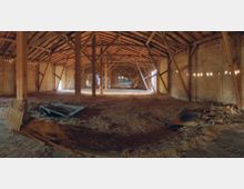 Innenansicht eines weitläufigen Scheunenraums mit Holzstützbalken und Holzdachkonstruktion. Der Boden ist mit Stroh bedeckt, und an den Wänden aus Backstein sind einige Lichtöffnungen sichtbar, die Sonnenlicht hereinlassen.