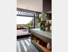 Eine moderne Terrasse mit Holzboden und gemütlichen Sitzbereichen, umgeben von Pflanzen. Im Vordergrund befindet sich eine offene Küchenzeile mit dekorativen Elementen wie Vasen und Pflanzen; im Hintergrund sieht man eine Pergola und eine städtische Aussicht.