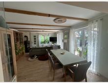 Offener Wohn- und Essbereich mit Holzbalkendecke, Esstisch mit Stühlen und angrenzender Sitzecke vor einem Kaminofen. Große bodentiefe Fenster mit hellen Vorhängen lassen viel Tageslicht herein und bieten Blick in den Garten; seitlich stehen Schränke und mehrere Zimmerpflanzen.