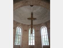Innenraum einer historischen Kirche mit gewölbter Backstein-Decke. Im Vordergrund hängt ein großes modernes Holzkreuz von der Decke, im Hintergrund sind drei hohe, schmale Fenster mit Rundbögen sichtbar.