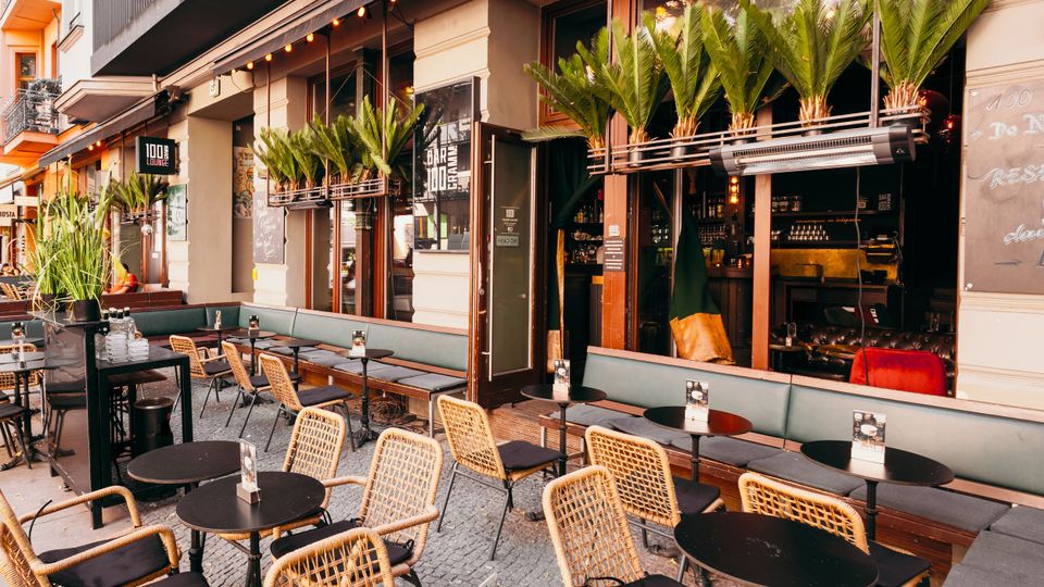 Außenbereich eines Straßencafés mit runden schwarzen Tischen, Korbstühlen und gepolsterten Sitzbänken entlang der Fassade. Große Fensterfronten mit offenen Türen, Pflanzkästen mit hohen Palmen und eine Lichterkette unter dem Vordach prägen die Szene.
