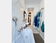 Schmales Badezimmer mit weiß gefliesten Wänden und einem weißen Fliesenboden. Im Vordergrund ist eine Badewanne mit Handbrause, daneben befinden sich ein Waschbecken und ein Handtuchheizkörper mit blauen und grünen Handtüchern. Im Hintergrund führt eine Türöffnung in einen anderen Raum.