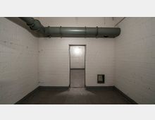 Ein kleiner Raum mit weißen Backsteinwänden, einem grauen Boden und einem großen Lüftungsrohr, das unter der Decke verläuft. In der Mitte befindet sich eine Türöffnung, die in einen weiteren, ähnlich gestalteten Raum führt; rechts unten ist ein kleines Belüftungsgitter sichtbar.