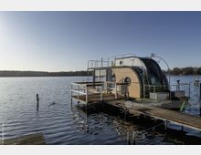 Modernes Hausboot mit Holzverkleidung und gewölbtem Glasdach, angedockt an einem Steg auf einem ruhigen See. Im Hintergrund ist eine bewaldete Uferlinie unter klarem, blauem Himmel sichtbar.