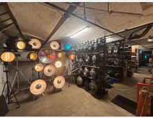 Raum mit industriellem Charakter und gewölbter Backsteindecke, gefüllt mit alten Film- und Bühnenscheinwerfern auf Ständern sowie Regalen, die mit weiteren Scheinwerfern und Equipment bestückt sind. Der Raum ist spärlich beleuchtet, wobei einige Scheinwerfer eingeschaltet sind und warmes Licht abstrahlen.