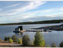 Schwimmendes Gebäude mit Vegetationsfassade auf einem See, verbunden mit einem kleinen Steg. Im Hintergrund ist eine von Bäumen gesäumte Uferlandschaft und ein gekrümmter Weg erkennbar.
