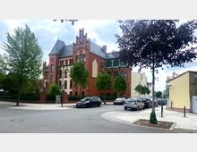 Historisches Backsteingebäude mit gotischen Elementen, wie Spitzbogenfenstern und Zinnen, umgeben von einer Straße und vereinzelten parkenden Autos; im Vordergrund und entlang der Straße sind Bäume zu sehen.