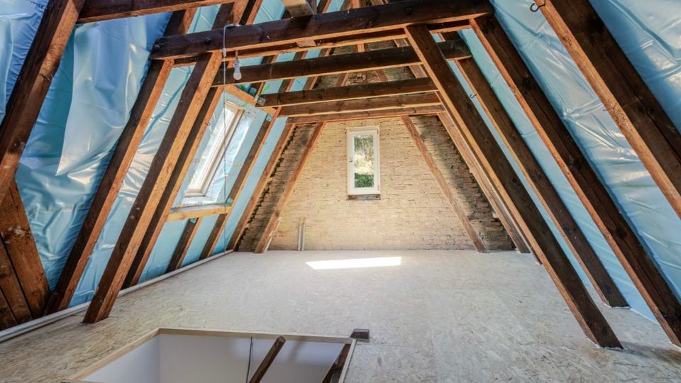 Ausgebauter Dachboden mit sichtbaren Holzbalken, gedämmten Dachschrägen mit blauer Folie und hellem Holzplattenboden. An der Stirnseite sind eine Ziegelwand sowie ein kleines Fenster und ein Dachfenster an der linken Schräge zu sehen.