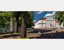 Weißes, barockes Gebäude mit rotem Walmdach, umgeben von Bäumen und einer gepflasterten Fläche mit vereinzelten Betonsitzen. Im Vordergrund stehen alte Bäume, während Informationsständer und eine Statue am Rand sichtbar sind.