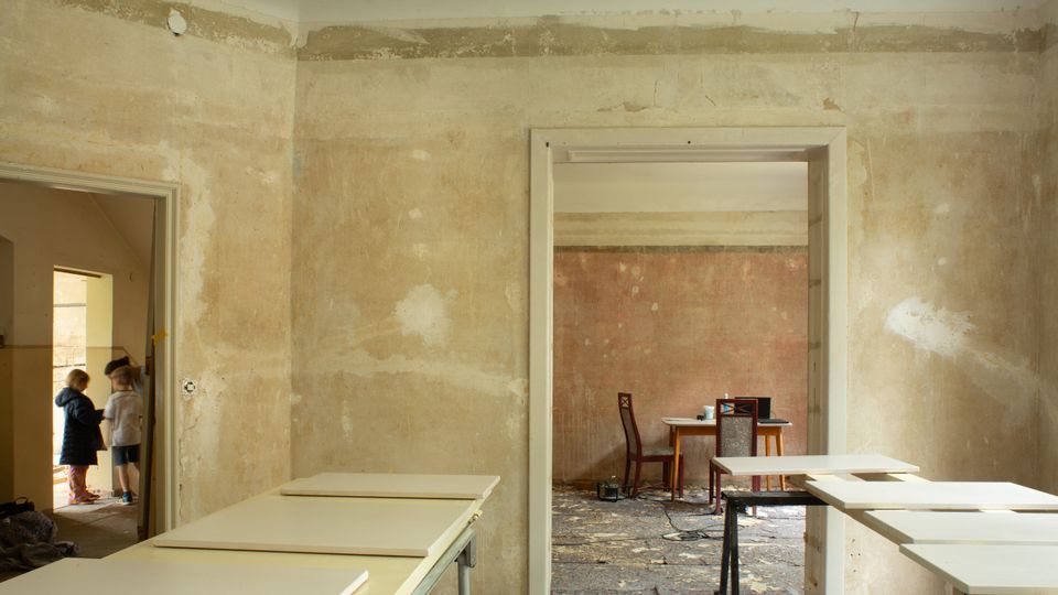 Innenansicht eines renovierungsbedürftigen Hauses mit roh verputzten, fleckigen Wänden und mehreren Türdurchgängen in angrenzende Räume. Im Vordergrund stehen provisorische Tische und Arbeitsplatten, im Hintergrund ein kleiner Tisch mit Stühlen auf einem staubigen Boden mit Bauschutt.
