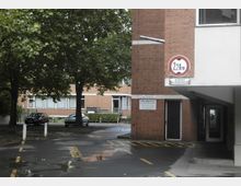 Backsteinfassade eines Gebäudekomplexes mit einem Durchgang und Hinweisschildern, darunter ein Höhenbegrenzungsschild (2,20 m). Im Hintergrund Parkfläche mit zwei geparkten Autos sowie Bäume und ein weiteres Gebäude mit großen Fenstern.