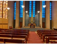 Innenraum einer modernen Kirche mit schlichten Holzbänken und einem zentralen roten Teppich, der zum Altarbereich führt. Die Wände sind mit mehreren Reihen blauer und gelber Buntglasfenster gestaltet, während der Altar auf einer erhöhten Plattform steht, flankiert von Kerzenständern.