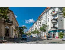 Straßenszene in einem Wohnviertel mit mehrstöckigen Altbauten, die weiße und beige Fassaden haben. Die Straße ist mit Kopfsteinpflaster bedeckt, gesäumt von geparkten Autos, kleinen Geschäften und Bäumen.
