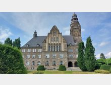 Historisches Gebäude aus Sandstein mit einem hohen Turm, der eine große Uhr zeigt, und einem steilen, mit Schiefer gedeckten Dach. Im Vordergrund befinden sich gepflegte Rasenflächen und hohe Bäume.