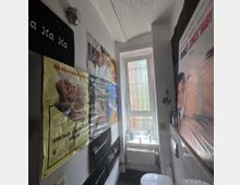 Schmales WC mit einem kleinen Fenster, durch das diffuses Tageslicht einfällt. Die Wände sind mit Filmplakaten bedeckt, und an der rechten Wand befinden sich weiße Fliesen, ein WC sowie ein kleines Heizkörper unter dem Fenster.