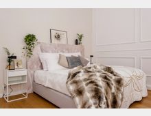 Schlafzimmer mit einem Doppelbett, das weiß bezogen ist und eine Bettdecke mit floralem Muster sowie ein Kunstfellplaid trägt. Ein helles Kopfteil aus gemustertem Material schmückt das Bett, während dekorative Kissen, Nachttische mit Pflanzen und minimalistische Wandkunst das moderne Design betonen.