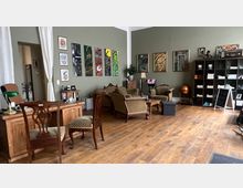 Ein stilvoll eingerichtetes Wohnzimmer oder Arbeitszimmer mit grünen Wänden, Holzboden und antik anmutenden Möbeln, darunter Sessel, ein Holzschreibtisch und ein Couchtisch. An der Wand hängen Gemälde, und ein Regal mit Büchern und Deko-Objekten steht rechts im Raum.