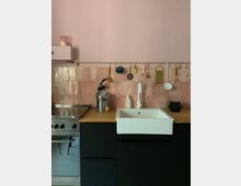 Kleine Küche mit rosa Wandfliesen als Rückwand, schwarzem Unterschrank, heller Holz-Arbeitsplatte und einem weißen Keramik-Spülbecken. An einer goldenen Stange über der Arbeitsfläche hängen Küchenutensilien, daneben befindet sich ein Herd aus Edelstahl.