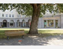 Städtischer Platz mit gepflastertem Weg und grünem Rasen, einer Holzbank und einem großen Baum im Vordergrund. Im Hintergrund befindet sich ein Gebäude mit Arkaden und mehreren Fenstern, darunter Geschäfte, darunter ein Geschäft mit dem Schriftzug "DAMENSALON".
