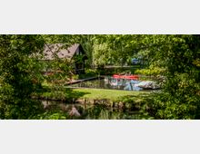Ein idyllischer Garten mit einem kleinen Teich, auf dem ein rotes Kanu mit vier Menschen zu sehen ist. Im Hintergrund steht ein Holzhaus mit Satteldach und umliegenden Bäumen, während Blumen und Rasenflächen den Teich umgeben.