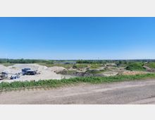 Eine offene Kiesgrube mit großen Sand- und Erdhaufen, Maschinen und Förderbändern im Vordergrund. Im Hintergrund sind grüne Landschaften, ein See, ein Windrad und ein weiter Himmel zu sehen.