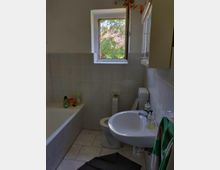 Kleines Badezimmer mit Badewanne, Waschbecken und Toilette. Ein Fenster mit Blick auf Bäume lässt Tageslicht herein, und an der Wand hängt ein grünes Handtuch.