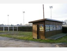 Holzhütte auf einem teilweise unbefestigten Gelände, umgeben von einer Hecke und weißem Zaun, mit mehreren Flutlichtmasten im Hintergrund. Werbeplakate sind an der Hütte angebracht, und im Hintergrund sind Häuser und ein offenes Feld sichtbar.