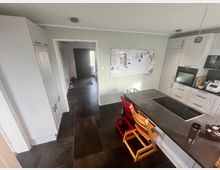 Moderne Küche mit einer Kochinsel, ausgestattet mit einem schwarzen Kochfeld. Weiße Einbauschränke und Elektrogeräte säumen den Raum. An der Wand hängt ein Whiteboard, und im Hintergrund führt ein offener Gang zu einer Haustür. Der Boden ist mit dunklen Fliesen ausgelegt.