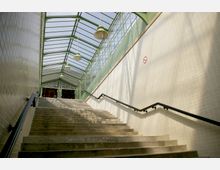 Treppe in einer überdachten Passage mit einem Glasdach und grünen Metallrahmen. Die Wände sind mit weißen Fliesen verkleidet, und an den Seiten befinden sich schwarze Handläufe.