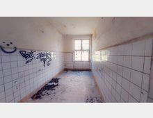 Ein schmaler, verlassener Raum mit weißen Fliesenwänden, teilweise beschmiert mit Graffiti. Am Boden sind Schutt und Blätter verteilt, und am Ende des Raums befindet sich ein einfaches Fenster, das nach draußen geöffnet ist.