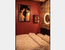 Schlafzimmer mit dunklen Ziegelwänden, aufgeteilt in eine rote und eine freiliegende Backsteinwand. Über dem Bett hängt ein großes Porträtbild, daneben ein kleiner Holzhängeschrank und ein runder, beleuchteter Spiegel. Das Bett ist mit floraler Bettwäsche bedeckt.