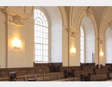 Innenraum einer historischen Kirche mit weißen Wänden, hohen Rundbogenfenstern, dekorativen Verzierungen und schlichten, hölzernen Kirchenbänken. An den Wänden befinden sich leuchtende Wandlampen.