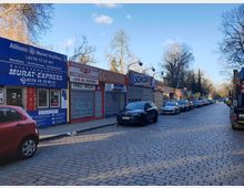 Reihe von kleinen Gewerbebetrieben mit bunten Schildern auf einer gepflasterten Straße; vor den Geschäften sind Autos entlang der Straße geparkt. Im Hintergrund sind Bäume zu sehen, die den Straßenrand säumen.