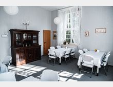 Ein heller Raum mit einfachen weißen Wänden, dekoriert mit einem großen Holzschrank im klassischen Stil. Mehrere Tische mit weißen Tischdecken und darauf platzierten Blumen stehen im Raum, umgeben von weißen Stühlen; durch ein großes Fenster mit durchscheinenden Gardinen fällt natürliches Licht herein.