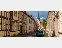 Schmale gepflasterte Straße in einer historischen Altstadt mit Fachwerkhäusern auf der linken Seite und verputzten Gebäuden auf der rechten Seite; im Hintergrund steht eine Kirche mit einem spitzen Turm und rotem Ziegeldach. Mehrere geparkte Autos säumen die Straße.