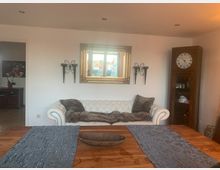 Ein Wohnzimmer mit weißem Ledersofa, dekoriert mit braunen Kissen und einer Decke. Über dem Sofa hängt ein goldgerahmter Spiegel, daneben ein hölzerner Schrank mit einer Wanduhr. Im Vordergrund steht ein Holztisch mit grauen Tischläufern.