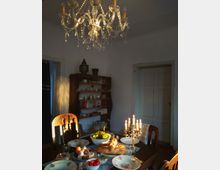 Esszimmer mit einem gedeckten Tisch, auf dem Brot, Obst und Kerzen auf einem goldfarbenen Kerzenhalter stehen. Im Hintergrund ein offenes Holzregal mit Geschirr und eine Kristall-Deckenleuchte, die den Raum beleuchtet.