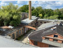 Blick über die Dächer eines älteren Industriekomplexes aus Backstein mit flachen, teils bitumengedeckten Dächern, Oberlichtern und einem hohen gemauerten Schornstein. Zwischen den Gebäuden liegen ein gepflasterter Hof und ein Roststeg, umgeben von dichtem Baumbestand und einem Gewässer im Hintergrund.