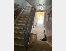 Treppenhaus in einem alten, renovierungsbedürftigen Gebäude mit einer Holztreppe, abgenutztem Geländer und Wänden mit abblätterndem Putz. Im Hintergrund ein lichtdurchfluteter Flur mit Holzboden.